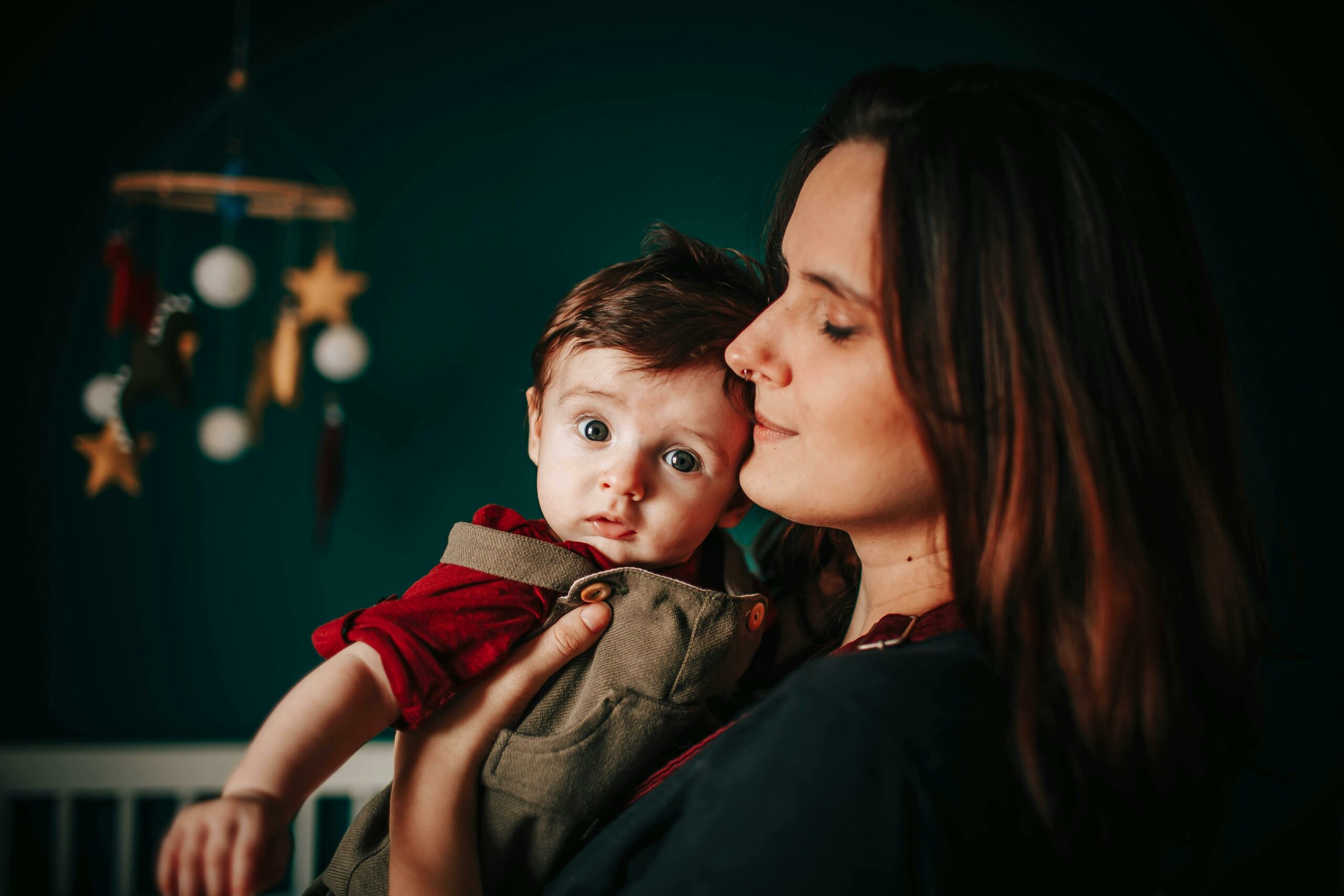 Caring mother with little son in arms standing in bedroom with white crib and baby mobile with toys at home