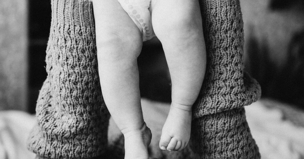 Tender moment of a mother kissing her baby's feet while lying in bed, captured in black and white.