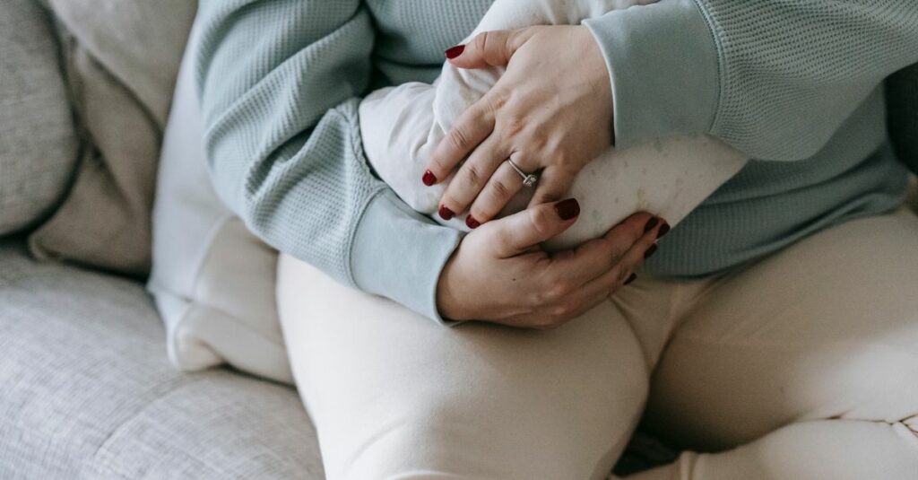 Crop caring faceless mother cuddling faceless newborn baby in arms resting on comfortable sofa in light living room at home